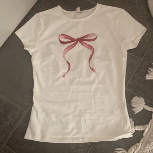 Vit t-shirt med rosett-tryck - Söt vit t-shirt med ett tryck av en rosa rosett framtill. Klassisk rund halsringning och korta ärmar. Perfekt för en enkel men gullig look.