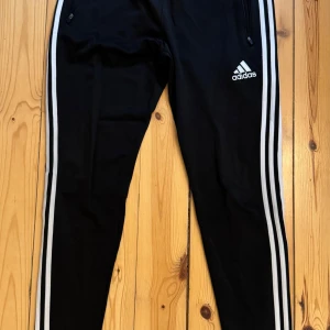 Svarta träningsbyxor från Adidas med vita ränder - Svarta träningsbyxor från Adidas med klassiska vita ränder längs sidorna och Adidas-logga på låret. Byxorna har dragkedjor vid fickorna och är tillverkade i ett lätt och svalt material med climacool-teknologi. Storlek M.