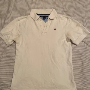 Vit piké t-shirt från Tommy Hilfiger - Klassisk vit piké t-shirt från Tommy Hilfiger med liten broderad logga på bröstet. T-shirten har korta ärmar, krage och knappar framtill. Perfekt för en stilren och avslappnad look.