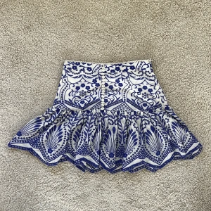 Vit och blå mönstrad kjol med volang - Supersöt kortkjol i vitt med blått broderat mönster och volang nertill. Kjolen har dekorativa knappar framtill 