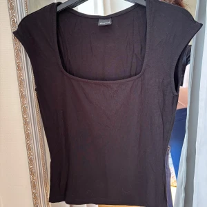 Svart topp med fyrkantig ringning från Gina! - En svart topp från Divided med fyrkantig ringning och korta ärmar. Toppen har en enkel och stilren design i mjukt bomullsmaterial. Perfekt att matcha med jeans eller kjol.