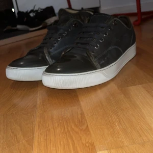 Svarta sneakers med vit sula från Lanvin - Säljer ett par svarta sneakers från Lanvin med vit, platt sula och rund tå. Skorna har snörning och är tillverkade i skinn med detaljer i syntet. Klassisk och stilren design som passar till mycket. Obs grön toe 