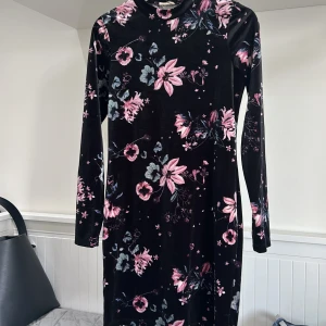 Svart sammetsklänning med blommigt mönster - Säljer en svart långärmad sammetsklänning med rosa och grått blommönster. Klänningen har en rak passform och hög hals, perfekt för dig som vill sticka ut med en mjuk och bekväm look.