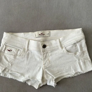 Vita jeansshorts från Hollister - Säljer ett par vita jeansshorts från Hollister med fransade benslut och slitna detaljer. Klassisk femficksmodell med låg midja och knappgylf. Perfekta för varma sommardagar!