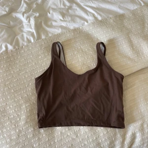 Brun croppad linnetopp från Gymshark - Säljer en brun croppad linnetopp från Gymshark med smala axelband och v-ringning. Toppen är i ett stretchigt material och har en liten logga framtill. Perfekt för träning eller som ett snyggt basplagg.