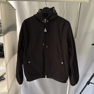 Svart vindjacka från Moncler med huva - Svart vindjacka från Moncler med huva och dragkedja framtill. Jackan har Moncler-logga på ärmen och detaljer i rött, vitt och blått på huvan. Två fickor med tryckknappar framtill och justerbar huva.