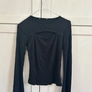 Svart ribbad långärmad topp med cut out - Säljer en svart ribbad långärmad topp med en snygg cut out-detalj vid bröstet. Materialet är stretchigt och sitter bekvämt på kroppen. Baktill är hel ribbat utan hål. 