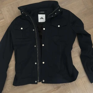 Blå jacka från J.Lindeberg - Stilren marinblå jacka från J.Lindeberg med silverfärgade knappar och dragkedja. Jackan är i storlek L och den är knappt använd.