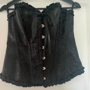 Svart korsett med spets och snörning - Säljer en svart korsett med vackert mönstrat tyg, volangkant upptill och nedtill samt snörning både fram och bak. Dekorativa metalldetaljer framtill och satinband för knytning. Perfekt för att skapa en snygg siluett.