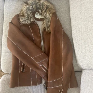 Brun jacka från Rino & Pelle - Säljer en brun jacka från Rino & Pelle i storlek 34 med fuskpäls runt kragen. Jackan har dragkedja framtill, dekorativa sömmar och två fickor med dragkedja. Priset går att diskutera💕