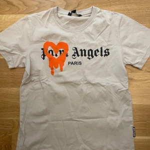 Beige t-shirt från Palm Angels - Säljer en beige t-shirt från Palm Angels med svart tryck och en orange hjärtformad detalj framtill. Klassisk passform och rund halsringning. Perfekt statement-plagg för dig som gillar streetwear.