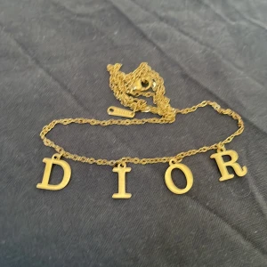 Guldigt halsband med DIOR-bokstäver - Säljer ett elegant halsband i guldton med kedja och hängande bokstäver som bildar ordet DIOR. Perfekt accessoar för att lyfta din outfit med en lyxig touch. Kedjan är tunn och smidig med klassisk länkdesign.