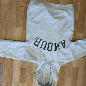 Ljusgrå hoodie med tryck från Gina Tricot - Säljer en ljusgrå hoodie från Gina Tricot med svart texttryck 'AMOUR' på framsidan. Tröjan har huva med dragsko och en klassisk känguruficka framtill. Perfekt för en avslappnad och trendig look.