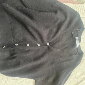 Svart kofta med glittriga knappar från Zara - Säljer en svart kofta från Zara med v-ringning och fina glittriga knappar i blomform. Koftan har lång ärm och är i ett mjukt, stickat material. Perfekt att slänga över en topp för en snygg look.