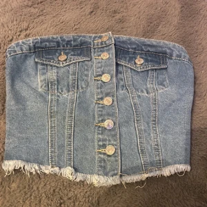 Jeans tubtopp med knappar och fransad kant - Säljer en snygg jeans tubtopp med knappar framtill och två bröstfickor. Toppen har fransad nederkant och smock baktill för bättre passform. Perfekt till sommaren och festivaler!