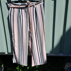 Randiga vida byxor med knytband - Säljer ett par ljusrosa byxor med svarta och vita vertikala ränder. Byxorna har vida ben och ett matchande knytband i midjan. Perfekta för dig som gillar en avslappnad men trendig stil.