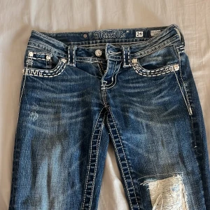 Miss Me bootcut jeans med broderade detaljer - Säljer ett par blå Miss Me jeans med bootcut passform köpte dom för 1500. Jeansen har vita broderade detaljer, dekorativa knappar och en patchad reva på ena benet. Snygga kontrastsömmar och detaljerade bakfickor med logga. Perfekta för dig som gillar statement-jeans.