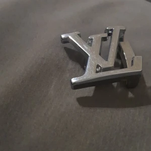 Louis Vuitton silverfärgat bältesspänne - Säljer ett stilrent bältesspänne från Louis Vuitton i silverfärgad metall. Spännet har den klassiska LV-loggan och texten 'Louis Vuitton' ingraverad. Perfekt för att ge din outfit en exklusiv touch.