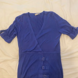 Blå omlottblus  - Snygg blå omlottblus med korta ärmar och ribbad midja. Blusen har två dekorativa spännen framtill och en v-ringad halsringning. 