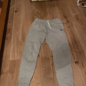 Grå mjukisbyxor från Reebok - Säljer ett par grå mjukisbyxor från Reebok med resår och snörning i midjan. Byxorna har smal passform och muddar vid bensluten. Perfekta för chill eller träning!