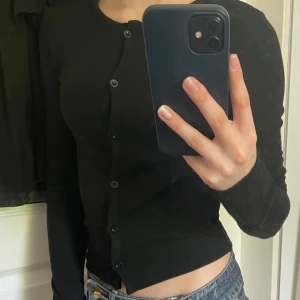 Svart kofta från Gina Tricot - Säljer en svart, tight kofta från Gina Tricot i storlek XS. Koftan har långa ärmar, rund halsringning och knäppning framtill med svarta knappar. Perfekt att styla till jeans eller kjol. Knappt använd