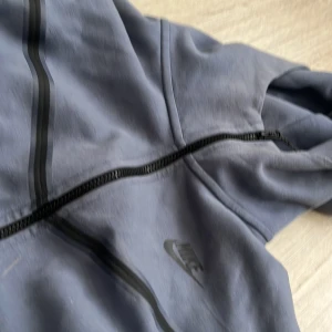 Blå zip-hoodie från Nike - Säljer en blå hoodie från Nike med dragkedja och huva. Hoddie har svarta detaljer längs dragkedjan och Nike-logga på bröstet. Perfekt för en sportig och avslappnad stil. Nike tech