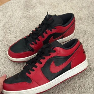 Nike Air Jordan 1 Low svart/röd - Säljer ett par Nike Air Jordan 1 Low i svart och rött med klassisk Jumpman-logga på plösen. Skorna har svart snörning, vit sula och röd yttersula. Perfekta för dig som gillar streetwear och sneakers med ikonisk design. Storlek 46! Använda fåtal gånger