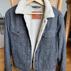 Grå jeansjacka med teddyfoder från Levi's - Säljer en grå jeansjacka från Levi's med vitt teddyfoder och klassisk krage. Jackan har två bröstfickor med knappar och stängs med knappar framtill. Perfekt för kyligare dagar.