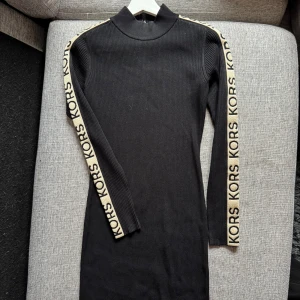 Svart ribbad klänning från Michael Kors - Svart ribbad långärmad klänning från Michael Kors med guld logoband längs ärmarna. Hög hals och figurnära passform. Perfekt för en stilren och trendig look.