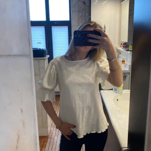 Vit blus med puffärmar - Säljer en söt vit blus med puffiga trekvartsärmar och lätt struktur i tyget. Blusen har en rund halsringning och är luftig i modellen, perfekt för vår och sommar.