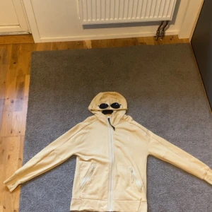 Beige zip Hoodie  med goggles från C.P. Company - Säljer en beige hoodiejacka från C.P. Company med ikoniska goggles i huvan. Jackan har dragkedja framtill och två fickor med dragkedja. Perfekt för dig som vill sticka ut med en unik streetwear-look.Den är äkta och i ny skick skriv vid intresse 