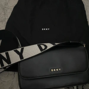 Svart axelväska från DKNY - Säljer en svart axelväska från DKNY med bred axelrem i svart och vitt med logga. Väskan har ett stilrent och minimalistiskt utseende med DKNY i guld framtill. Perfekt för att bära det viktigaste.