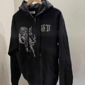 Svart hoodie med ängel och strasstenar - Säljer en svart hoodie med dragkedja från BV. På framsidan finns en stor ängel och BV-logga i strasstenar. Tröjan har huva och långa ärmar, perfekt för en cool och avslappnad stil.