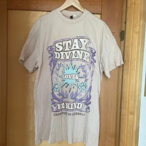 Beige t-shirt med tryck från Divided - Beige t-shirt från Divided med stort retroinspirerat tryck i lila, blått och vitt framtill. Texten 'STAY DIVINE', 'LOVE', 'BE KIND' och blommor ger en cool vintage vibe. Rund halsringning och korta ärmar. Perfekt för en avslappnad stil. Har lite rosa fläckar från tt ha hamnat i färgad tvätt