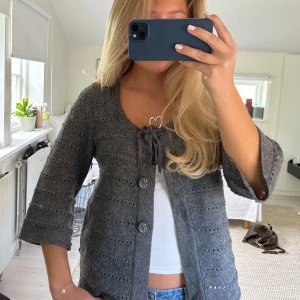 Grå stickad kofta med knytband - Grå stickad kofta med trekvartsärm och stora knappar framtill. Söt knytning vid halsen och diskret hålmönster i materialet. Perfekt att slänga över en topp för en chill look.