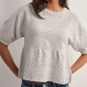 Ljusgrå stickad topp med volang - Supermjuk ljusgrå stickad topp med korta ärmar och volang nedtill. Rund halsringning och avslappnad passform. Perfekt att styla med jeans för en chill look.