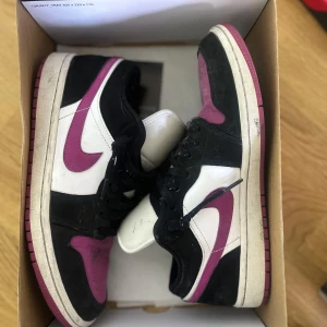 Nike Air Jordans Low svart/vit/lila - Säljer ett par Nike Air Jordans Low med svart, vit och lila färgkombination. Skorna har klassisk swoosh i lila på sidorna, svart ovandel och vit mittpanel. Yttersulan är lila och de har snörning framtill. Perfekta för dig som gillar streetwear och sneakers.