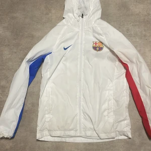 Vit FC Barcelona vindjacka från Nike - Säljer en vit vindjacka från Nike med FC Barcelona-logga på bröstet. Jackan har huva, dragkedja och fickor. Ena ärmen har blå detalj och den andra röd, vilket ger en sportig look. Perfekt för fotbollsfans!