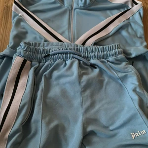 Ljusblå mjukisbyxor från Palm Angels - Säljer ett par ljusblå mjukisbyxor från Palm Angels med vita och svarta ränder längs sidorna. Byxorna har resår i midjan och är tillverkade i ett mjukt syntetmaterial. Perfekta för en avslappnad stil.