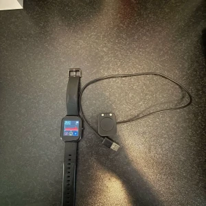Svart smartklocka med laddare - Säljer en svart smartklocka med fyrkantig urtavla och silikonarmband. Klockan har touchskärm och flera funktioner, samt medföljer en magnetisk USB-laddare. Perfekt för dig som vill ha koll på notiser och hälsa direkt på handleden.Klockans urarmband har gåt sönder tyvär.
