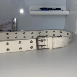 Vitt bälte med dubbla spännen - Säljer ett vitt bälte i canvas med dubbla metallspännen och silverfärgade öljetter längs hela bältet. Perfekt för att ge outfiten en cool touch. Bältet har en robust känsla och passar till många olika stilar.