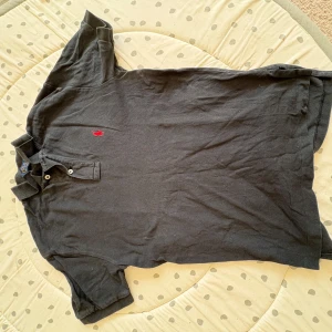 Svart pikétröja från Polo Ralph Lauren - Klassisk svart pikétröja från Polo Ralph Lauren med röd broderad logga på bröstet. Tröjan har korta ärmar, krage och knappar vid halsen. Tillverkad i bomull för en bekväm känsla. Storlek XS väldigt bra skick