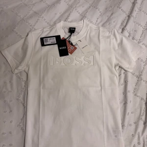 BOSS T-shirt - Säljer en stilren vit t-shirt från BOSS med upphöjd logotyp framtill. Klassisk passform och rund halsringning. Perfekt för en clean och modern look. (Sitter som M)