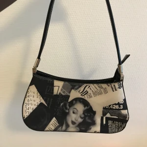 Svartvit axelväska med tidningsmönster - Trendig svartvit axelväska med coolt tidningsinspirerat mönster och retrobild. Väskan har en smal axelrem och dragkedja upptill. Perfekt för dig som vill sticka ut med en unik accessoar.