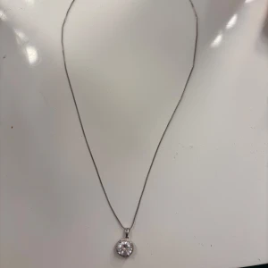 Silverhalsband med rund glittrig sten från SNO - Säljer ett elegant silverhalsband från SNO med en rund, glittrig sten som hänge. Kedjan är tunn och smidig, perfekt för att ge en stilren touch till din outfit. Hänget har en gnistrande sten omgiven av små detaljer.