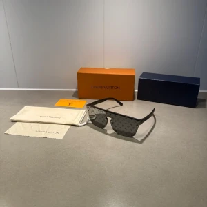 Svarta LV glasögon  - Snygga svarta solglasögon från Louis Vuitton med LV-monogram på glasen. Rektangulär form och breda skalmar i svart plast. Kommer med originalfodral, putsduk och ask. Perfekt för dig som vill sticka ut med en exklusiv accessoar.