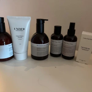 Hud- och hårvårdsprodukter från Under Your Skin och Revolution - Flera produkter för hud och hår, inklusive en vit tub med fuktbehandling, bruna pumpflaskor och sprayflaskor samt en hår-olja i kartong. Märkena är Under Your Skin och Revolution. Förpackningarna är stilrena i vitt och brunt.