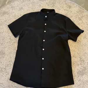Svart kortärmad skjorta från SHEIN - Säljer en stilren svart skjorta från SHEIN i storlek S. Skjortan har korta ärmar, klassisk krage och vita knappar framtill. Perfekt för en clean och enkel look. Materialet känns lätt och bekvämt.
