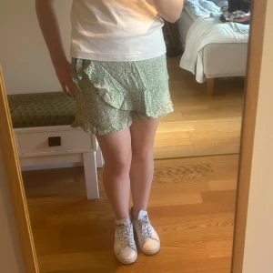 Grön blommig kjol med inbyggda shorts - Säljer en grön kjol med inbyggda shorts med vitt blommönster. De har omlottdesign med knyt i midjan och volangdetaljer längs kanterna. Perfekta för varma dagar och ger en söt och somrig look.