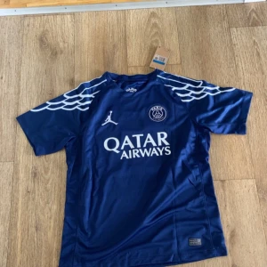 PSG fotbollströja O. Dembélé 10 - Mörkblå PSG fotbollströja med vita detaljer, Jordan-logga och Qatar Airways sponsortryck. Vingar på axlarna och O. Dembélé 10 på ryggen. Helt ny,först till kvarn